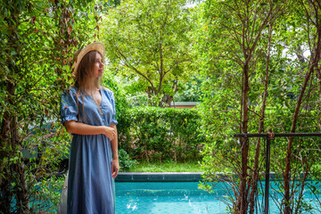 A woman in a stylish straw hat strolls by a crystal-clear swimming pool set amidst tropical...