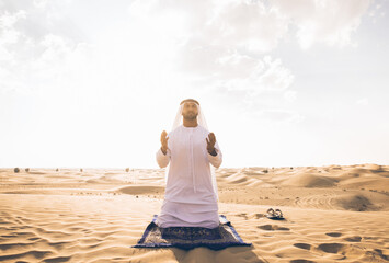 Man with white traditional kandura from uae praying in the desert on the carpet