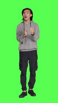 Front view of asian happy guy applauding a person, clapping hands to show his congratulations and cheering for someone over full body greenscreen. Young man feeling optimistic and cheerful, standing