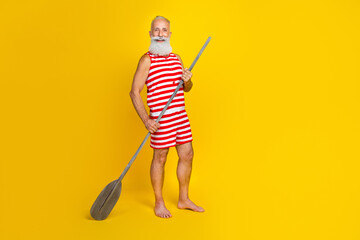 Full body size photo of professional experienced old sportsman standing on boat holding wooden oar isolated on yellow color background