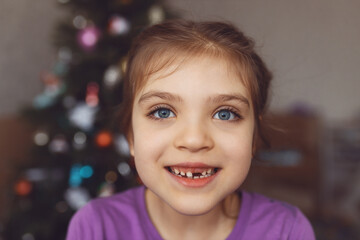 Funny girl 6 years old without one front top tooth against background of Christmas tree at home