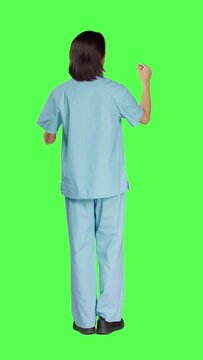 Back view of asian medical nurse checks hologram in studio with greenscreen design, working with holographic icon tab and artificial intelligence. Male specialist in uniform with healthcare expertise.