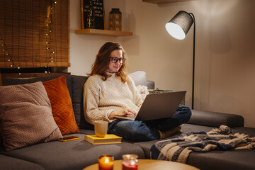 A young woman spends a cozy winter evening at home lying on the sofa with a laptop. Winter holidays, Christmas and online surfing