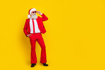 Full length photo of cool handsome man pensioner dressed red suit santa hat looking empty space isolated yellow color background - obrazy, fototapety, plakaty