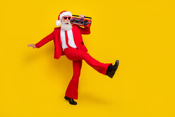 Full length photo of pretty funky man pensioner dressed red suit santa hat walking listening boom box isolated yellow color background