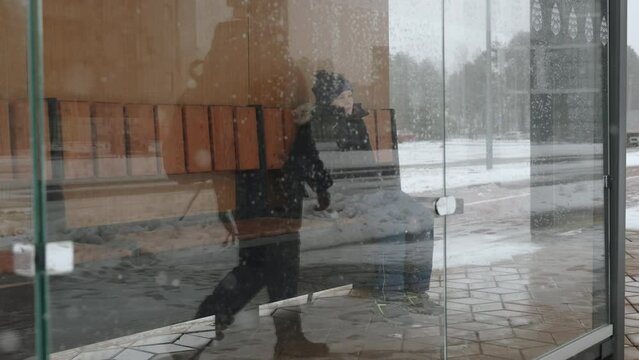 Woman sits down near lonely boy on bench at public transport stop, view behind glass. Persons looks around indifferently. Inattention to other people problems. Respecting child personal boundaries.