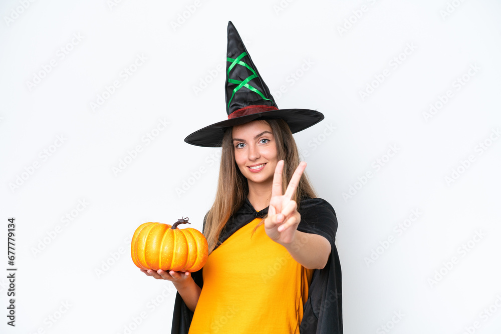 Wall mural Young caucasian woman costume as witch holding a pumpkin isolated on white background smiling and showing victory sign