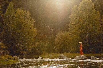 Fototapeta premium Woman with straw hat in middle of forest with river in background trees and nature warm colors, meditating in nature, traveling through east