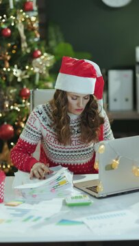 Christmas time. tired elegant middle aged business woman in santa hat and red Christmas sweater with documents and laptop working in modern green office with Christmas tree.