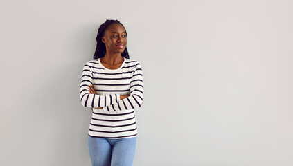 Happy beautiful young slim African American woman in casual clothes standing with crossed arms by a light gray copy space wall background, looking away and smiling - obrazy, fototapety, plakaty