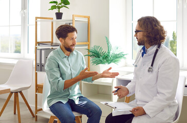 Young man patient talking with male doctor in his office in clinic during medical examination. Male physician listening to patient's complaints making appointment. Healthcare and medicine concept.