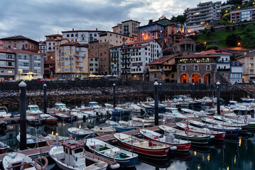 Fototapeta na wymiar View of the old town of Mutriku village beginning to light up at dusk with the fishing port full of boats in the foreground, Gipuzkoa, Basque Country, Spain