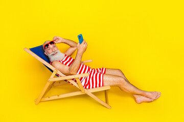Full length photo of carefree old man lying on sunbed at turkish resort using smartphone and relaxing isolated on yellow color background