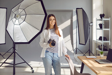 Young woman photographer with professional camera in photo studio. Cheerful young woman holding professional dslr camera standing in office with professional photographic equipment
