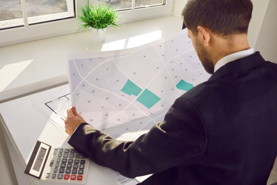Male professional engineer analyzing cadastral map in office. Shoulder view shot of cartographer sitting at desk working with printed map, evaluating, accounting cost and locating building plot