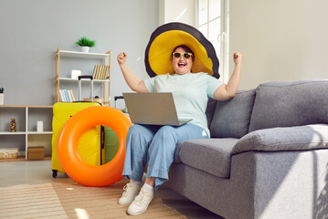 Funny fat plus size woman in summer sunhat and sunglasses sitting on sofa with laptop, suitcases...