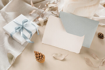 Congratulation blank paper card, envelope, gift box with blue ribbon, fir cones on beige background.