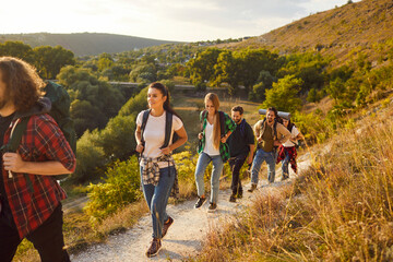 Active wanderlust people going hiking or trekking on good sunny day. Group of male female friends with backpacks following mountain trail route, leaving countryside with green woods and valleys behind - obrazy, fototapety, plakaty