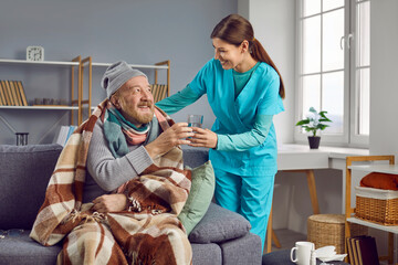 Smiling nurse caring about elderly unwell patient at home. Female caregiver in uniform giving glass...