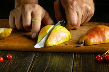 Close-up of a man hands on a kitchen table using a knife to cut a fresh pear on a wooden board to prepare dessert
