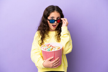 Obraz premium Little caucasian girl isolated on purple background surprised with 3d glasses and holding a big bucket of popcorns