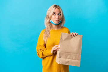 Young Russian woman holding a grocery shopping bag isolated on blue background having doubts while...