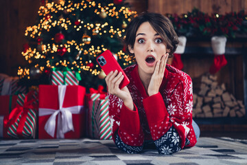 Portrait of impressed cute person lying floor carpet hand touch cheek use smart phone giftbox tree garland indoors