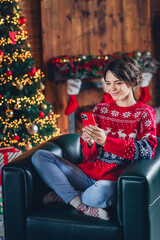 Full length vertical photo of peaceful person sit comfy chair use smart phone x-mas shopping garland lights house indoors