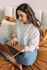 woman with long curly hair in light clothes works online from home. Remote work at the computer. Girl freelancer studies online at home sitting on the bed