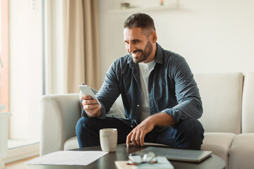 Middle aged man smiles texting via cellphone in living room