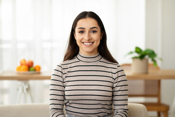 Headshot of happy young pretty arab woman looking at camera in light living room interior