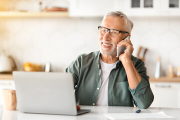 Smiling Elderly Gentleman Using Laptop And Talking On Cellphone In Kitchen