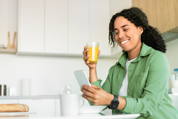 black woman looking at phone, drinking fresh orange juice indoor - obrazy, fototapety, plakaty