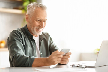 Happy Older Gentleman Using Smartphone While Sitting At Desk In Kitchen