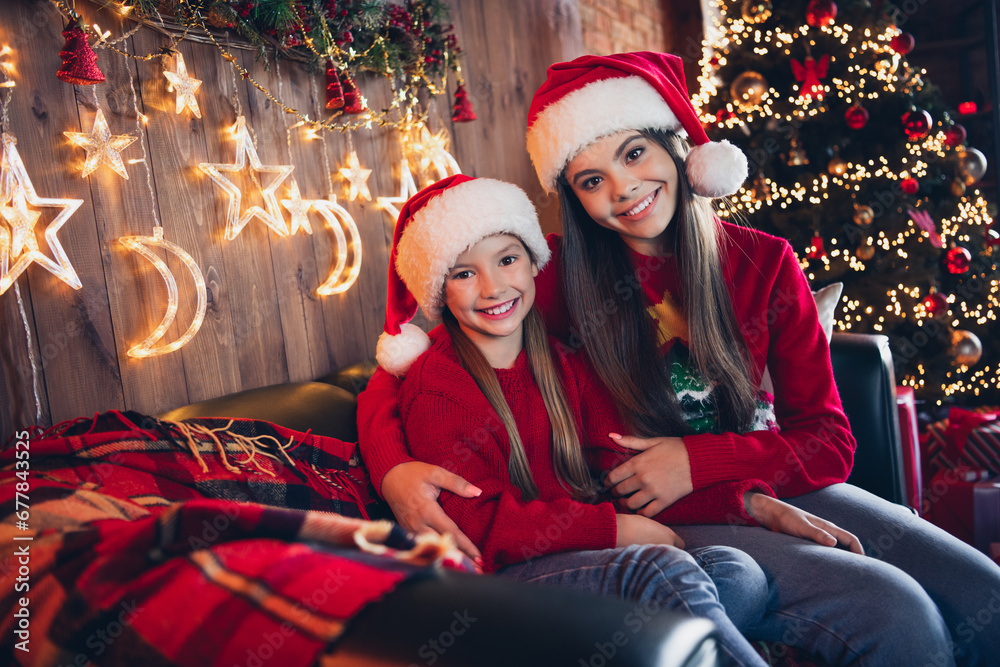 Poster Photo of dreamy pretty small sisters wear red ugly print new year sweaters hugging embracing indoors apartment room