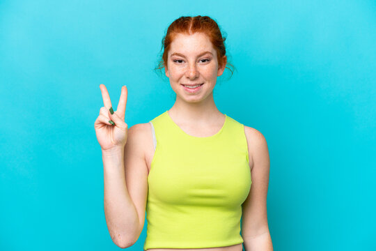 Young reddish woman isolated on blue background smiling and showing victory sign