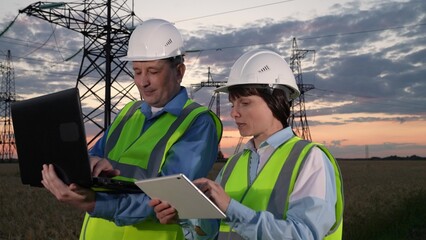 Engineers compare information on computers by power transmission lines at sunset