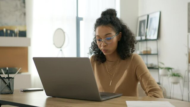 Education people slow. Smart multi ethnical freelancer woman in eyeglasses working on computer at home. Casual concentrated curly female entrepreneur developing new project working on laptop inside 4K