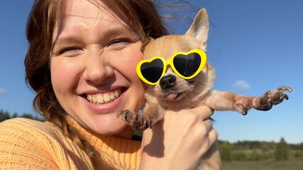 Happy young woman or teenager girl walking with her puppy Chihuahua dog in funny sunglasses in the forest or park at sunny day - obrazy, fototapety, plakaty