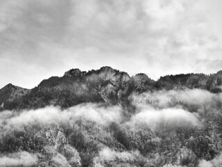 Majestic and rugged Swiss alps show through a mass of thick and puffy clouds, in black and white.

