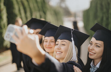 University students celebrate graduation in a sunny park. Smiling, they take selfies, and share...