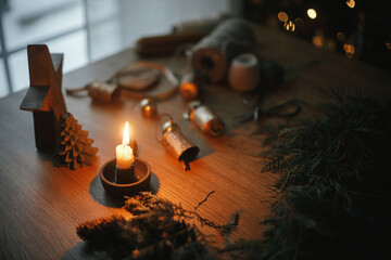 Candle, cedar branches, pine cones, twine, bells on wooden table in evening festive room. Making...