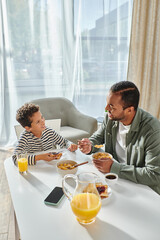 vertical shot of joyful african american father and son enjoying breakfast and smiling happily