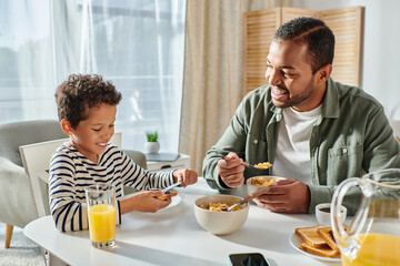 Fototapeta na wymiar cheerful handsome african american father looking happily at his son eating breakfast at table
