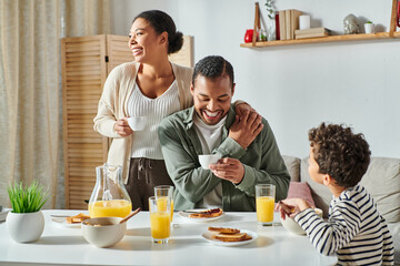 cheerful african american parents with coffee cups in hands smiling near their son eating breakfast