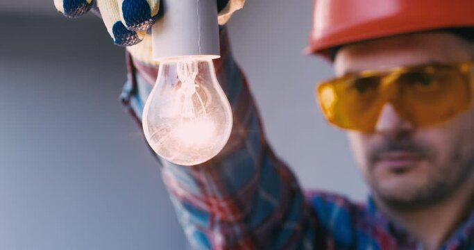 The electrician scraews the light bulb into the socket with a gloved hand, the light bulb begins to glow.