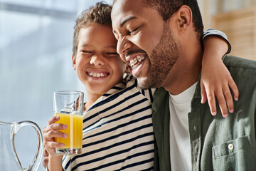 joyous african american father and son smiling happily with closed eyes, orange juice in hand