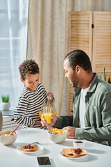 vertical shot of cute little african american boy pouring glass of orange juice with his father help