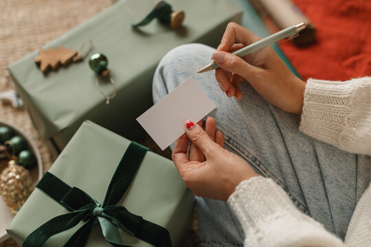 Close-up of a woman sitting cross-legged on the floor next to a wrapped Christmas gift writing a Christmas card