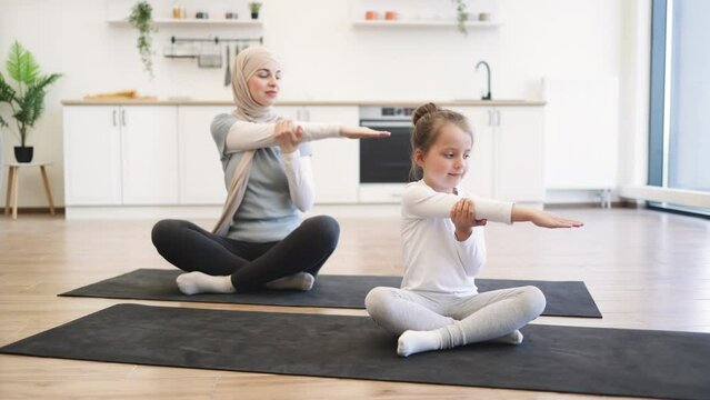 Front view of small female kid on yoga mat stretching arms with slim muslim lady with closed eyes during training. Young parent and daughter in sportswear spend time happily during workout.